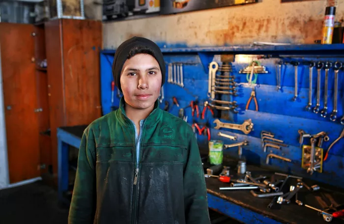 A boy in a green work jacket and black beanie stands confidently in a workshop surrounded by tools neatly arranged on a blue wall.