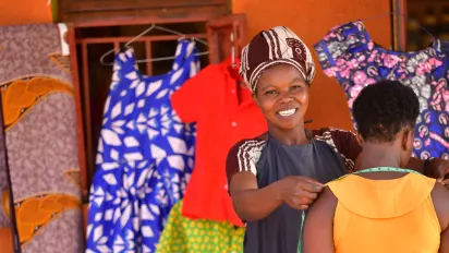 A female tailor smiles at the camera while she takes measurements of another woman's shoulders