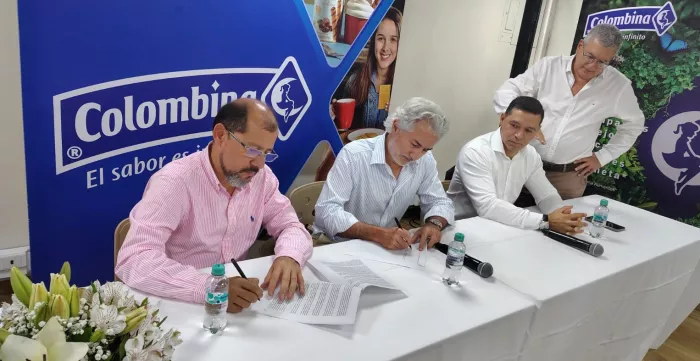 Tres hombres sentados, uno de ellos firma un documento. Al fondo un banner de la empresa Colombina